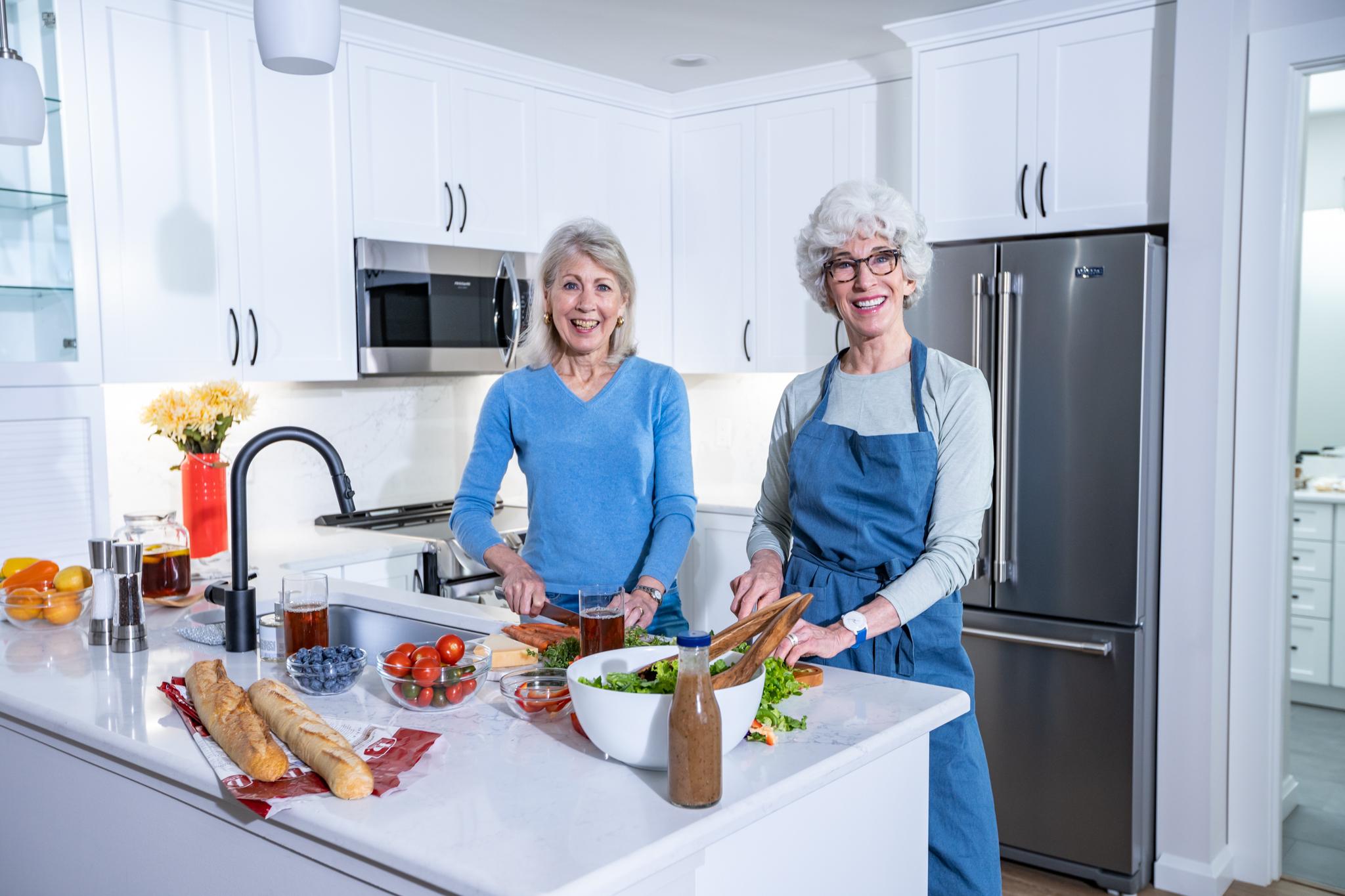 Two older women smile as they look up from preparing a healthy meal.