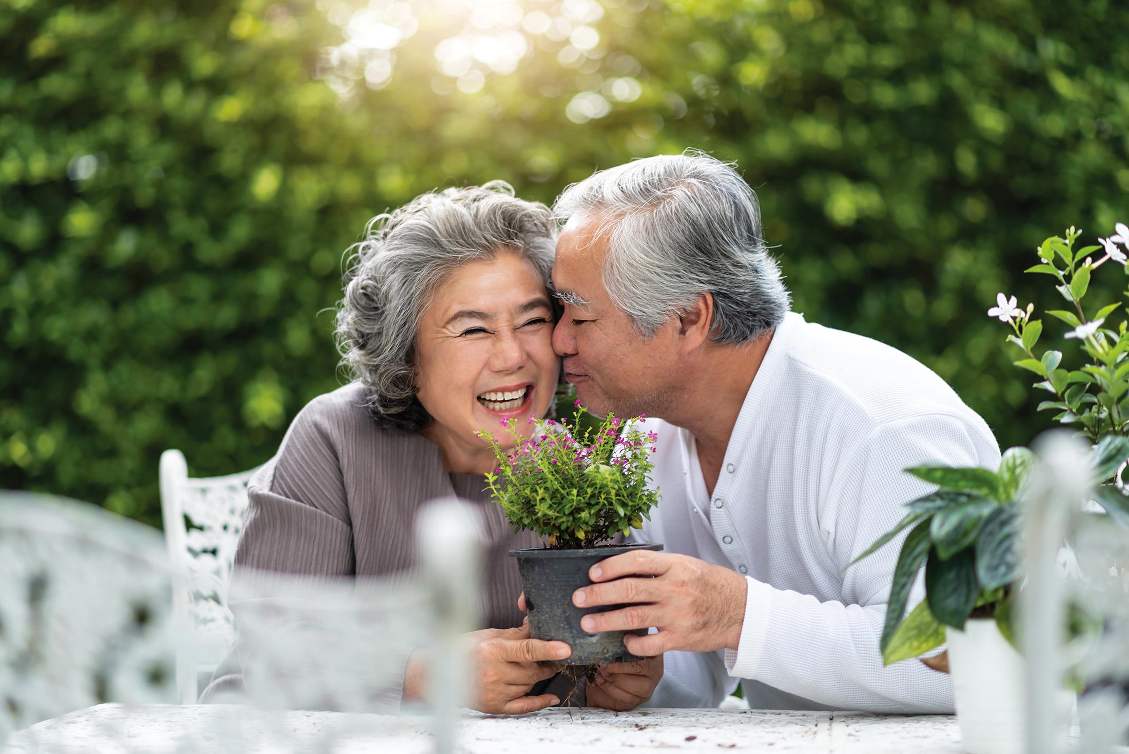 An older man and his wife smile and embrace outdoors while holding a potted plant.