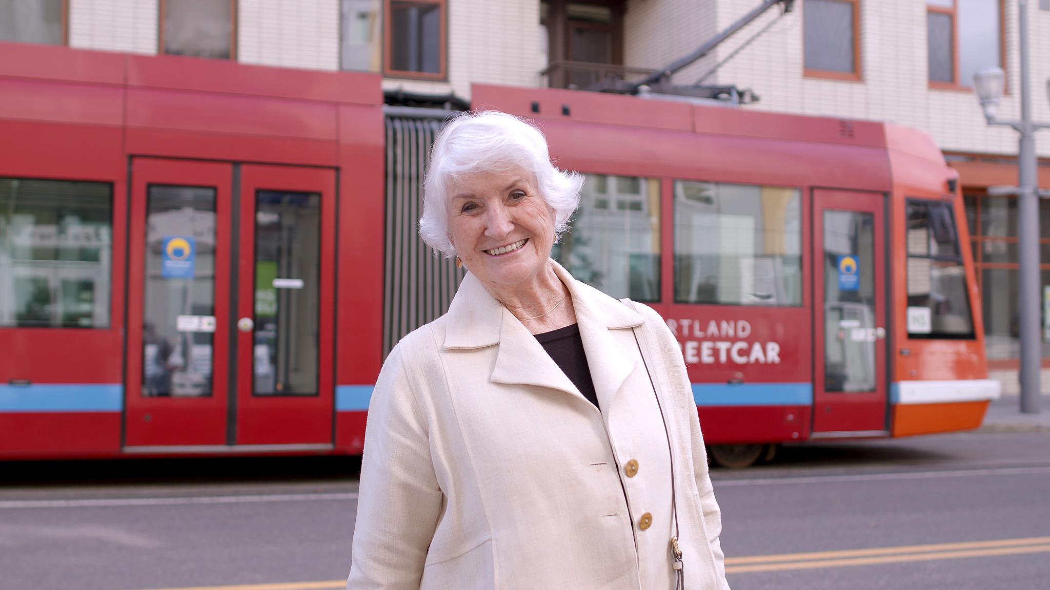 An older woman smiles for the camera in front of a streetcar.