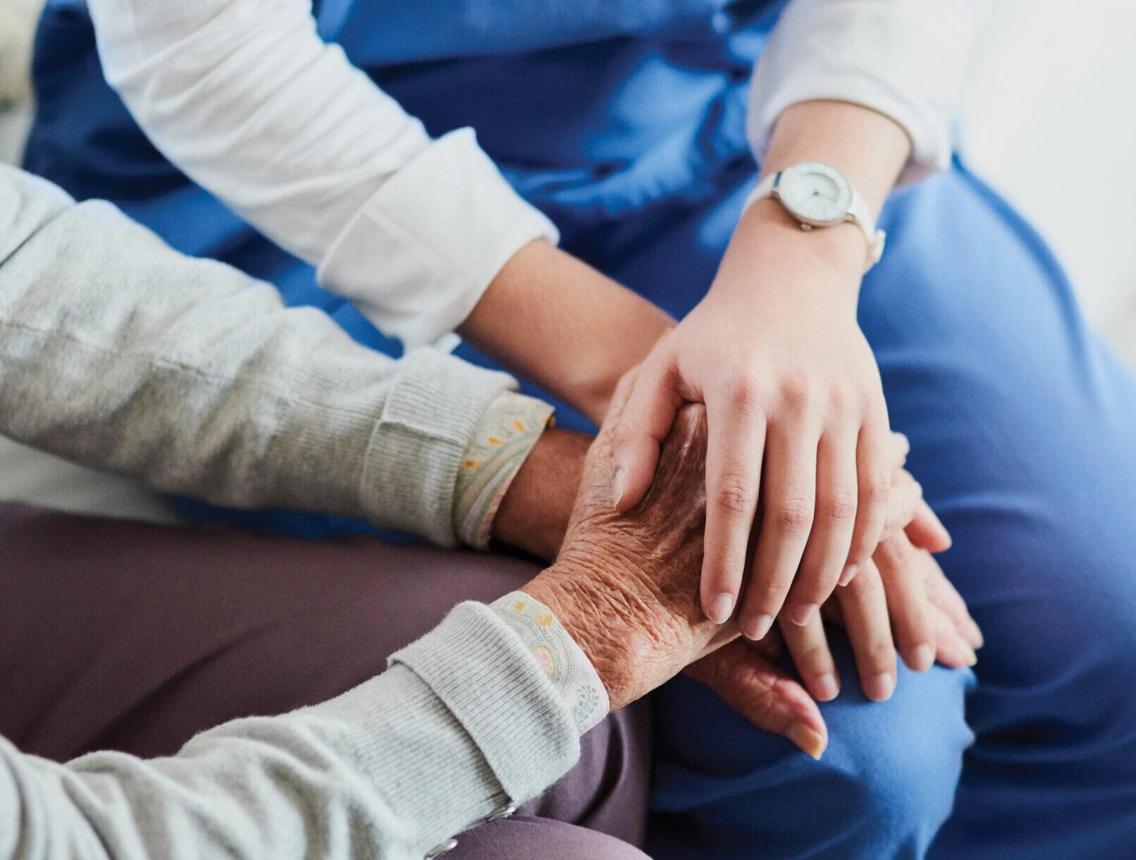 A compassionate nurse holds a senior patient’s hands. Their heads are out of frame.