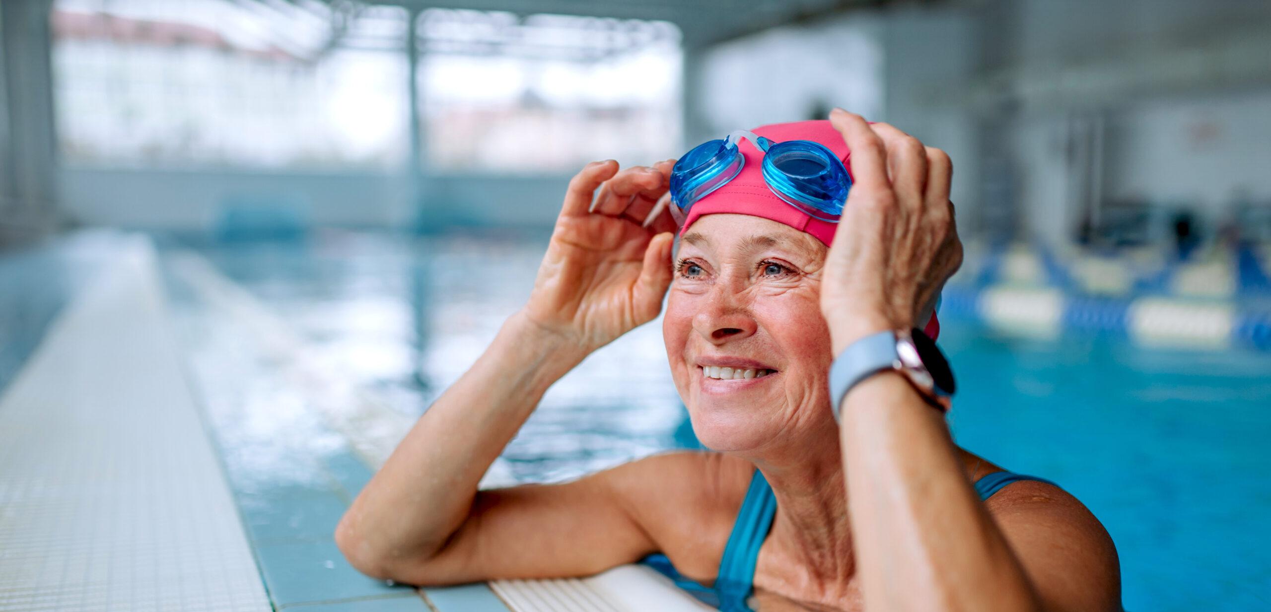 A happy senior woman leans against the side of the pool and adjusts her goggles.